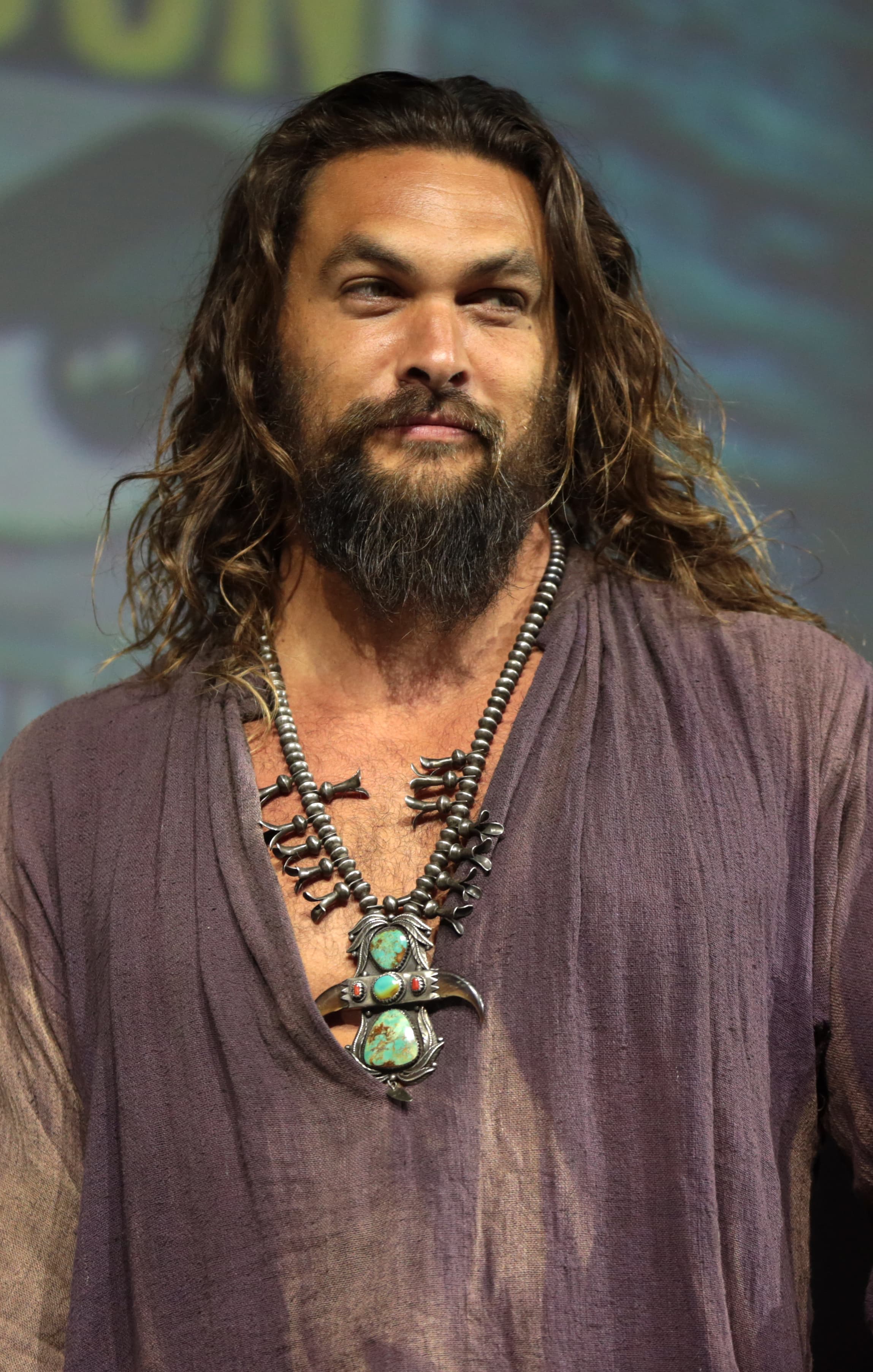 Jason Momoa at San Diego Comic-Con in 2018 wearing a purple shirt and necklaces